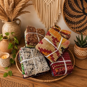 May include: A collection of folded fabric squares featuring various patterns and colors, including brown, yellow, red, and black and white, all tied together with white string and arranged in a round woven basket on a wooden surface.