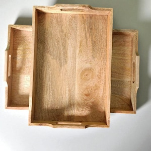 May include: Three rectangular wooden serving trays with built-in handles. The trays are made of light-colored wood, showcasing the natural grain. The trays are empty and ready for use.