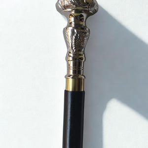 Può includere: Un bastone da passeggio color argento con un motivo floreale decorativo sull'impugnatura e un fusto in legno marrone scuro. Il bastone ha un collare in ottone e un intarsio color oro vicino al fondo.