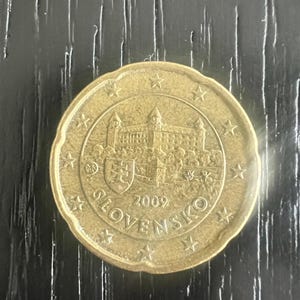 May include: A gold-coloured coin with a detailed castle design, the year 2009, and the word "SLOVENSKO". The coin has a scalloped edge and is set against a dark, textured background.