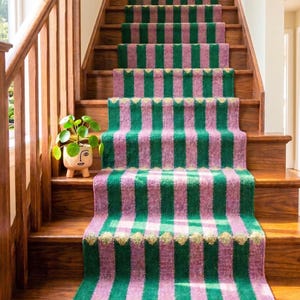 May include: A stair runner featuring vertical stripes in alternating shades of forest green and dusty pink. The runner includes small, repeating gold-coloured diamond accents across the width and is finished with a fringe at the bottom edge.