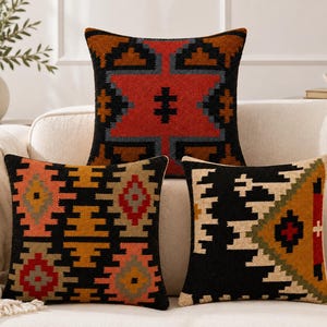 May include: Three square throw pillows featuring intricate woven geometric patterns in black, red, orange, yellow, and cream. The top pillow displays a large central star motif, while the two bottom pillows feature repeating diamond and triangular shapes.