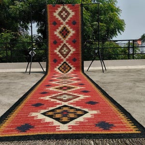 Puede incluir: Una alfombra pasillera larga y rectangular con una base de color rojo vibrante y un patrón geométrico repetitivo de diamantes en negro, blanco y tostado. La alfombra tiene un borde negro sólido y flecos cortos claros en el extremo.