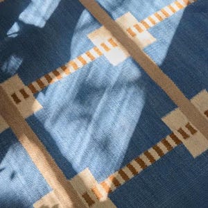 May include: A woven textile featuring a blue background with a geometric grid pattern. The design consists of intersecting tan lines and light cream rectangular accents at the junctions, creating a structured, repeating grid across the fabric surface.