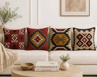May include: Four square throw pillows arranged on a white sofa. The first is red with black geometric patterns. The second is red with a central diamond motif in yellow and black. The third features a yellow and green hexagon design. The fourth is beige with black and red diamond shapes.