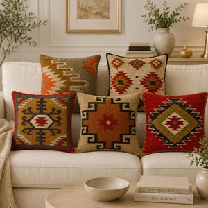May include: Five square throw pillows featuring woven geometric patterns in shades of red, orange, yellow, brown, and cream. Each pillow displays a distinct symmetrical design with intricate lines and shapes, arranged on a light-colored sofa.