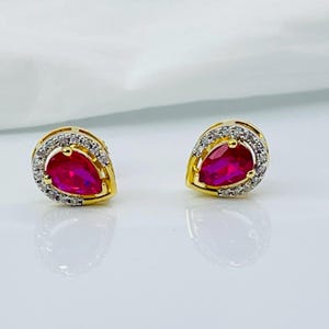 Könnte beinhalten: Ein Paar goldfarbene Ohrstecker. Jeder Ohrring hat einen tropfenförmigen, leuchtend roten Edelstein, der von einem Kranz aus kleinen, klaren Steinen umgeben ist. Die Ohrringe sind vor einem weißen Hintergrund platziert, der das Licht reflektiert.