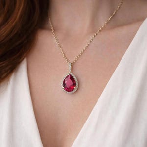Peut inclure: Un collier en or avec un pendentif en forme de goutte d'eau. Le pendentif présente une grosse pierre rouge vif entourée d'un halo de petites pierres claires et scintillantes. Le collier est porté sur un fond blanc.