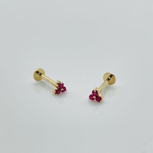 Puede incluir: Dos pendientes de botón dorados, cada uno con un grupo de tres pequeñas piedras preciosas rojas redondas. Los pendientes tienen una parte trasera plana y circular y un poste recto. El fondo es claro.