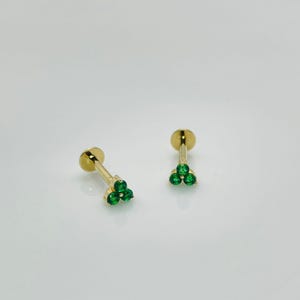 Könnte beinhalten: Zwei goldfarbene Ohrstecker, jeweils mit einem Cluster aus drei leuchtend grünen Edelsteinen. Die Ohrringe haben eine flache, runde Rückseite und einen schlanken Steg. Die Edelsteine sind in einem dreieckigen Muster angeordnet.