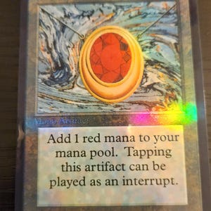 Pu&ograve; includere: Carta di Magic: The Gathering con un ciondolo d'oro con una grande gemma rossa. La carta ha un effetto olografico e il testo "Add 1 red mana to your mana pool."