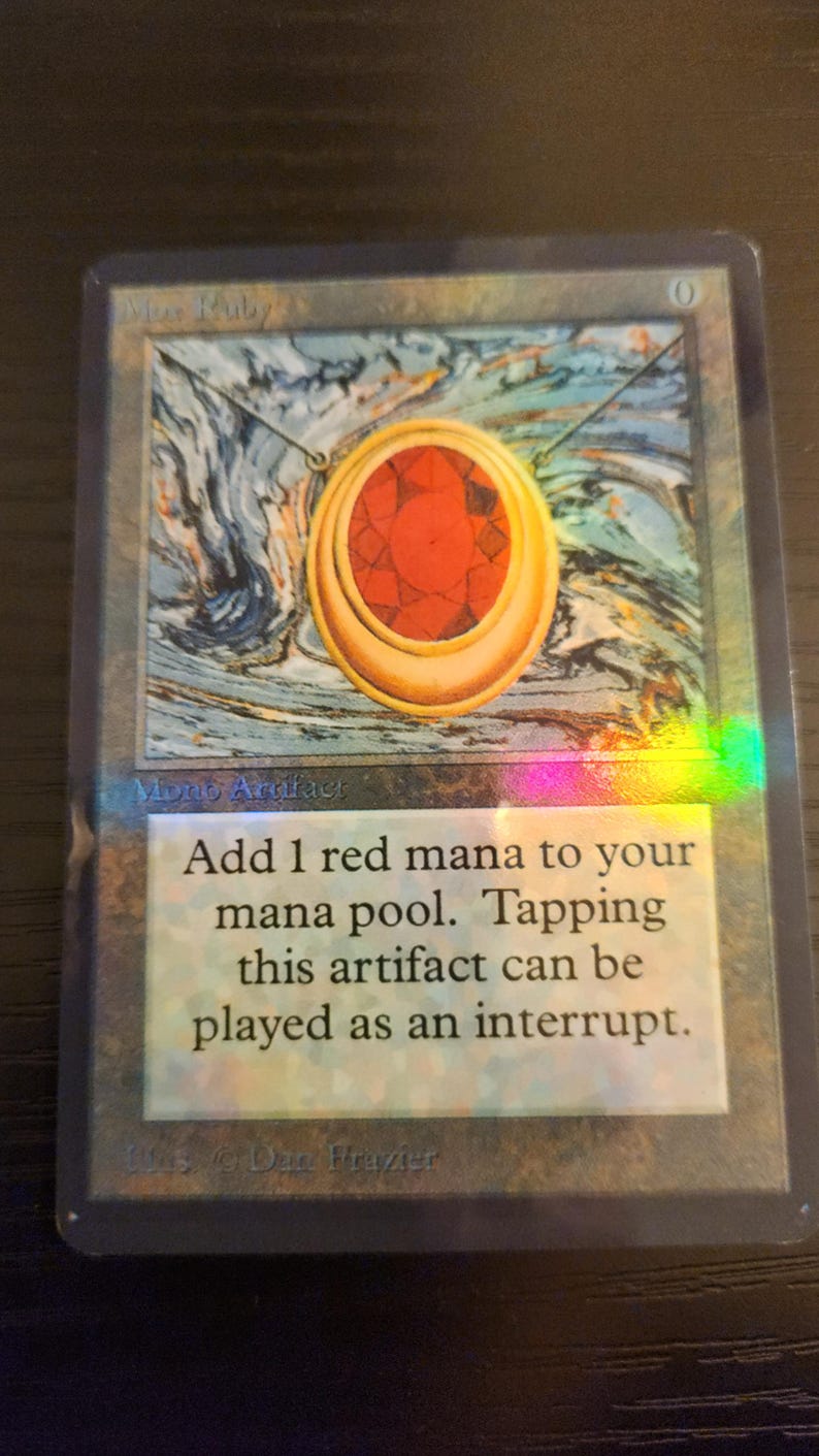 Pu&ograve; includere: Carta di Magic: The Gathering con un ciondolo d'oro con una grande gemma rossa. La carta ha un effetto olografico e il testo "Add 1 red mana to your mana pool."