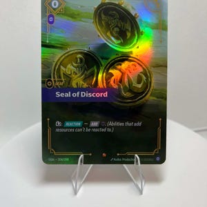 May include: A holographic trading card featuring three gold coin illustrations with a central emblem. The card reads "Seal of Discord" and includes game instructions. The card is displayed on a clear stand.