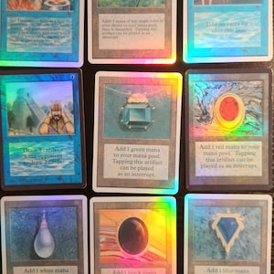 May include: Nine collectible trading cards with holographic foil accents. The cards feature various fantasy illustrations and text, including a blue lotus flower, a green gem, and a red orb. The cards are for a collectible card game.