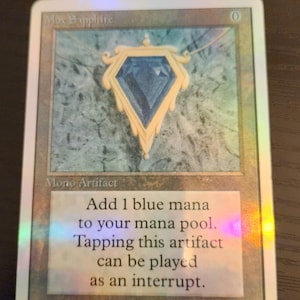 Pu&ograve; includere: Una carta collezionabile con un ciondolo a forma di gemma blu con dettagli dorati. La carta recita "Mox Sapphire" e "Mono Artifact". Il testo sulla carta spiega la sua funzione nel gioco. &Egrave; visibile anche il nome dell'artista, Dan Frazier.