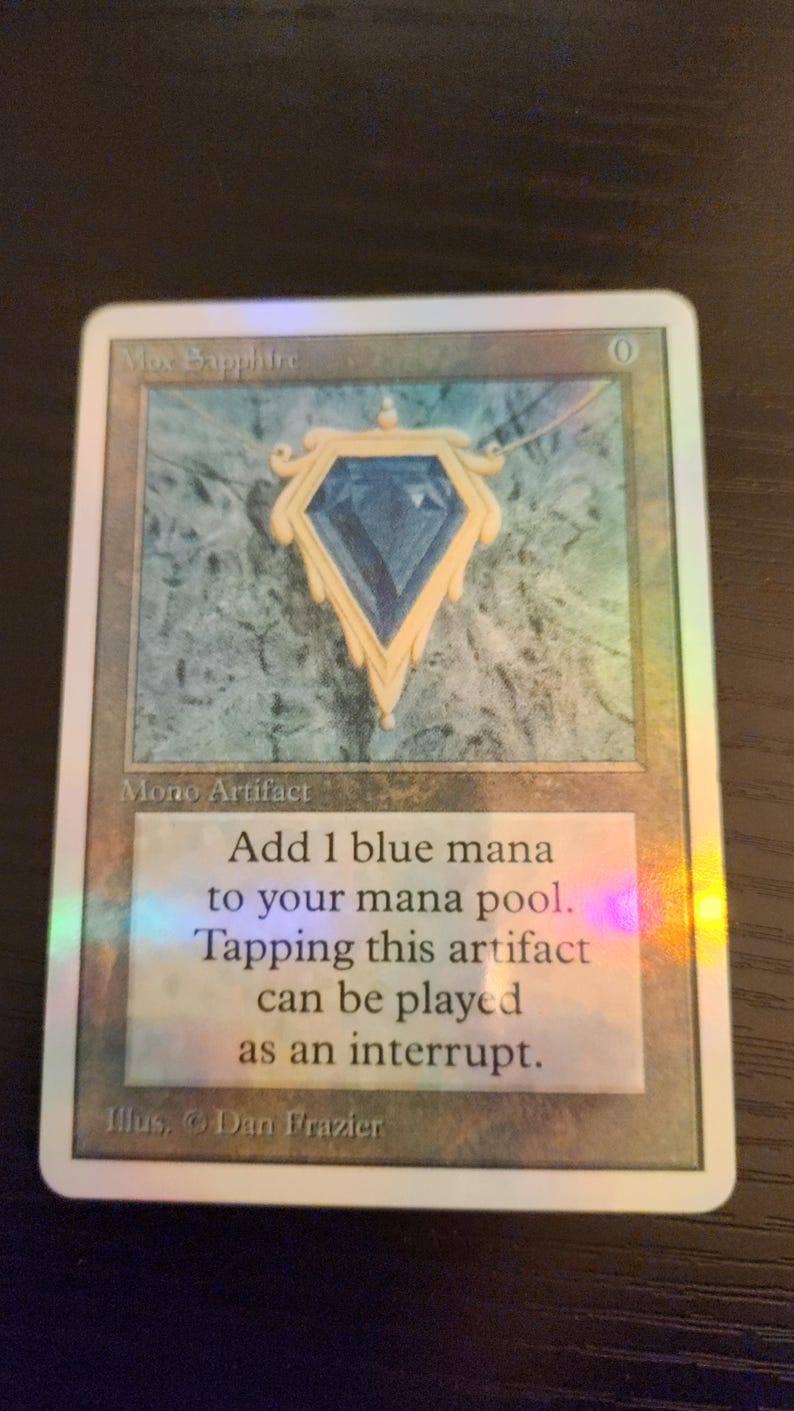 Pu&ograve; includere: Una carta collezionabile con un ciondolo a forma di gemma blu con dettagli dorati. La carta recita "Mox Sapphire" e "Mono Artifact". Il testo sulla carta spiega la sua funzione nel gioco. &Egrave; visibile anche il nome dell'artista, Dan Frazier.