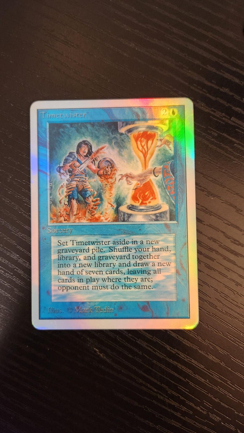 Pu&ograve; includere: Una carta olografica di Magic: The Gathering intitolata "Timetwister". La carta presenta un'illustrazione di una figura e una clessidra infuocata. Il testo descrive la funzione della carta come magia.