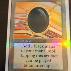 Pu&ograve; includere: Una carta di Magic: The Gathering con un artefatto circolare nero, una catena d'oro e uno sfondo colorato. Il testo della carta recita: "Aggiungi 1 mana nero al tuo mana pool. Tappare questo artefatto pu&ograve; essere giocato come interruzione."