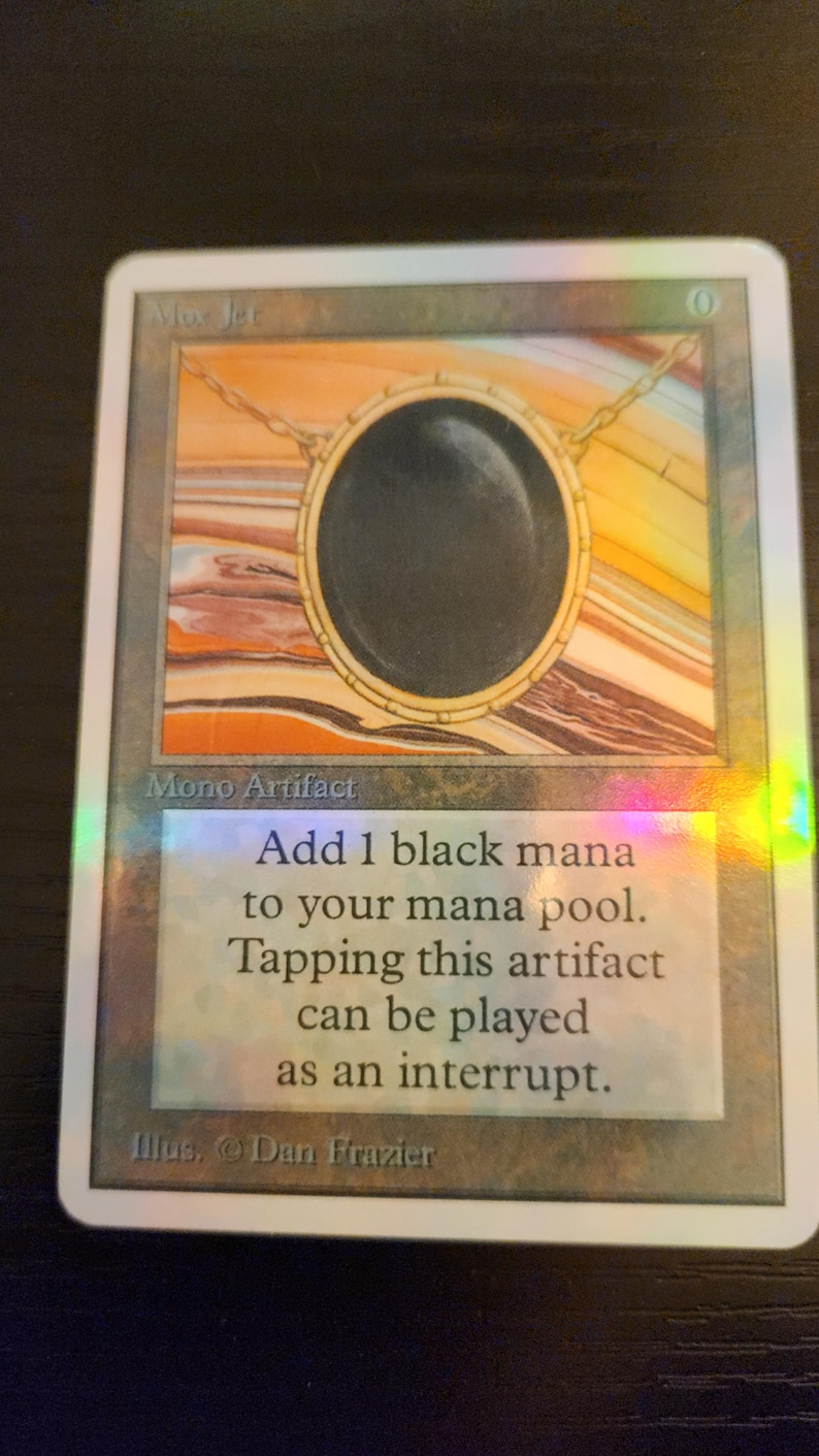 Pu&ograve; includere: Una carta di Magic: The Gathering con un artefatto circolare nero, una catena d'oro e uno sfondo colorato. Il testo della carta recita: "Aggiungi 1 mana nero al tuo mana pool. Tappare questo artefatto pu&ograve; essere giocato come interruzione."