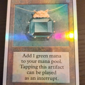 Pu&ograve; includere: Una carta di Magic: The Gathering con un ciondolo di gemma verde-azzurro con chiusura dorata. La carta &egrave; intitolata "Mox Emerald" e presenta testo che descrive la sua funzione nel gioco. Illustrazione di Dan Frazier. Carta rettangolare.