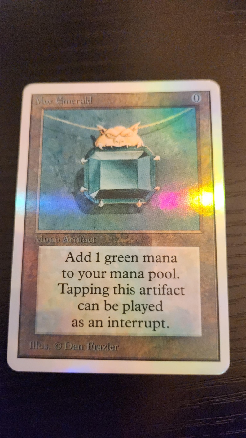 Pu&ograve; includere: Una carta di Magic: The Gathering con un ciondolo di gemma verde-azzurro con chiusura dorata. La carta &egrave; intitolata "Mox Emerald" e presenta testo che descrive la sua funzione nel gioco. Illustrazione di Dan Frazier. Carta rettangolare.