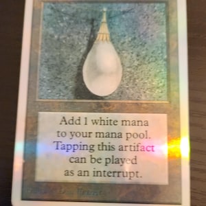 Pu&ograve; includere: Carta olografica di Magic: The Gathering con un oggetto bianco a forma di goccia. Il testo della carta recita: "Aggiungi 1 mana bianco al tuo mana pool. Tappare questo artefatto pu&ograve; essere giocato come interruzione."