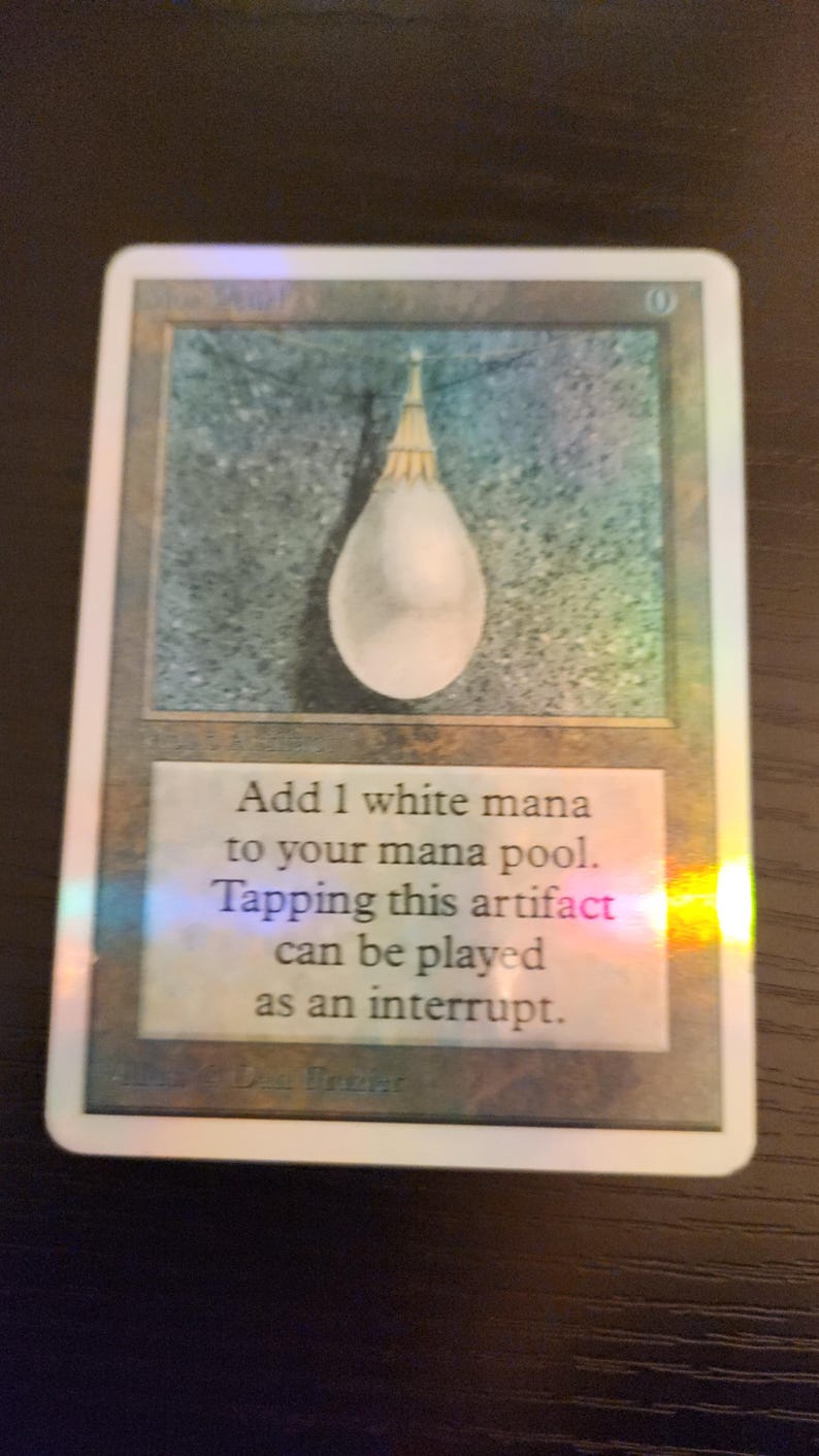 Pu&ograve; includere: Carta olografica di Magic: The Gathering con un oggetto bianco a forma di goccia. Il testo della carta recita: "Aggiungi 1 mana bianco al tuo mana pool. Tappare questo artefatto pu&ograve; essere giocato come interruzione."