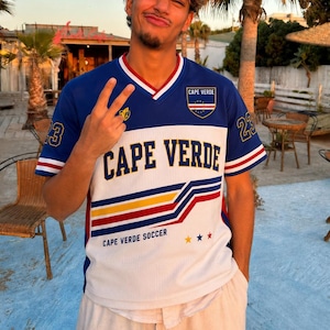 Op de afbeelding: Een blauw en wit voetbalshirt met V-hals en de tekst CAPE VERDE in dikke marineblauwe letters op de borst. Het shirt bevat een embleem van Kaapverdië, het nummer 23 op de mouwen, rode en gele horizontale strepen en CAPE VERDE SOCCER tekst.