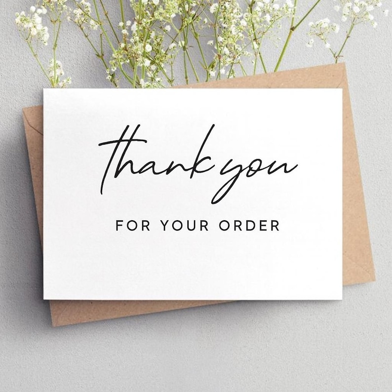May include: A white card with the words "thank you" in a handwritten style and "for your order" in a simple font. The card is on a brown envelope, with white flowers in the background.