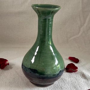 May include: A ceramic vase with a glossy finish, featuring a bulbous base that tapers into a slender neck and a flared rim. The upper portion is a vibrant green, transitioning into a deep, dark purple hue at the rounded bottom section.
