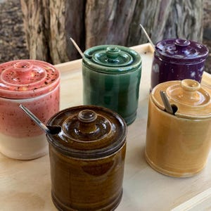 Puede incluir: Conjunto de cinco tarros de cerámica en varios colores, incluyendo rosa, verde, morado, marrón y amarillo. Cada tarro tiene una tapa y una pequeña cuchara. Los tarros están colocados sobre una bandeja de madera.