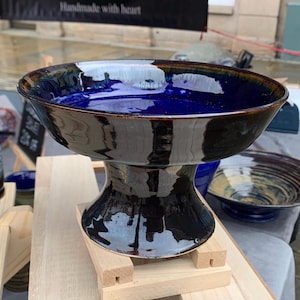 Puede incluir: Un cuenco con pedestal de cerámica hecho a mano, con un interior azul oscuro brillante y un exterior negro. El cuenco tiene una base ancha y poco profunda y una base acampanada. El borde tiene un acento marrón. Se exhibe sobre un soporte de madera.