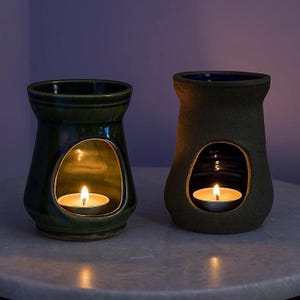 May include: Two ceramic oil warmers, one dark green and one textured brown, each with an oval opening and a lit tea light candle inside. The warmers are on a light-coloured surface.