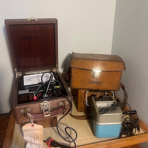 May include: A wooden box with a handle, meter, and wires, sits on a wooden tray. A leather case with "MEGGER" is next to a silver and blue device. A tag hangs from the wooden box. The items appear to be electrical testing equipment.