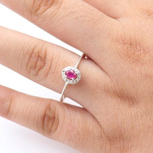 May include: A silver ring featuring a round, bright red gemstone surrounded by a halo of smaller, clear stones. The ring is set on a thin band and is displayed on a hand with light green nail polish.