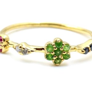 Puede incluir: Un delicado anillo de oro con un diseño floral y varias piedras preciosas. El anillo tiene una banda delgada y está adornado con un grupo de piedras verdes, rojas y negras. El anillo se presenta sobre un fondo blanco.