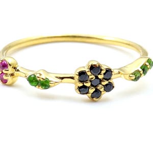 Peut inclure: Une bague en or délicate avec un motif floral. La bague a un anneau fin et est ornée d'un groupe de pierres précieuses noires au centre, flanqué d'accents de pierres précieuses roses et vertes, ressemblant à des fleurs et des feuilles.