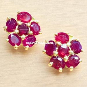 Può includere: Un paio di orecchini a bottone dorati, ciascuno con un gruppo di pietre preziose rosse scure di taglio ovale. Le pietre sono disposte a motivo floreale, creando un design scintillante ed elegante. Gli orecchini sono su uno sfondo chiaro.