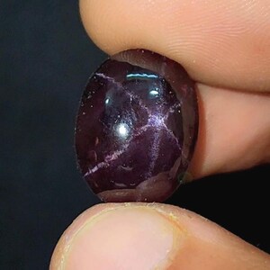 May include: A deep purple gemstone with a star-like pattern. The oval-shaped stone is held between two fingers, highlighting its polished surface and internal light reflections. The gem's rich colour and unique pattern make it a striking piece.
