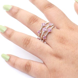 Könnte beinhalten: Ein silberner Ring mit drei Bändern, die jeweils mit kleinen, quadratischen, rosafarbenen Edelsteinen besetzt sind. Der Ring wird an einem Finger getragen und zeigt das detaillierte Design und das Funkeln der Edelsteine. Der Ring ist ein Schmuckstück.