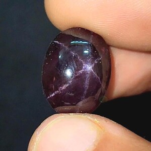 May include: A deep purple, oval gemstone with a polished finish. The stone displays a star-shaped pattern of lighter inclusions. The gem is held between fingers, highlighting its smooth, reflective surface. The gem is likely a cabochon.