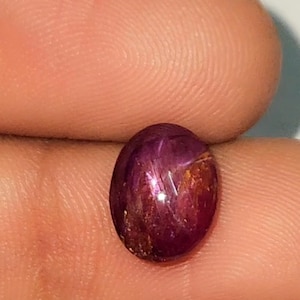 Può includere: Una gemma di rubino lucidata, di forma ovale. La gemma è di un rosso intenso con sfumature viola e dorate. La superficie riflette la luce, creando un effetto stellato. Il rubino è tenuto tra le dita.