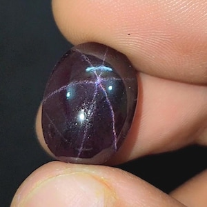 May include: A deep purple, oval gemstone with a polished surface. The stone displays a star-shaped pattern of intersecting lines. The gem is held between fingers, highlighting its form and the play of light across its surface.