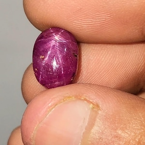 May include: A polished, oval ruby gemstone. The gem is a deep red with a star effect across its surface. The ruby is held between fingers, showcasing its colour and lustre.
