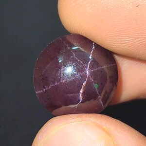 May include: A polished, round gemstone in a deep burgundy hue. The stone features a star-shaped pattern of light lines across its surface. The gemstone is held between two fingers against a dark backdrop.
