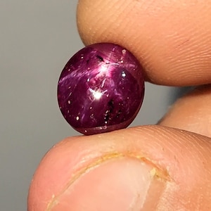 May include: A polished, round ruby gemstone, held between fingers. The gem is a deep red with a star-like effect, and small white inclusions. The surface reflects light, creating a brilliant shine. The gemstone is a cabochon cut.