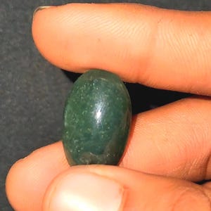 May include: A polished, oval-shaped green gemstone. The stone is held between two fingers, showcasing its smooth, reflective surface. The gemstone's color is a deep, rich green, with subtle variations in tone.