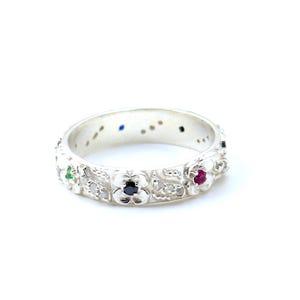 Peut inclure: Une bague en argent avec un motif floral. La bague est ornée de petites pierres précieuses colorées, dont du vert, du rouge, du bleu et du noir. La bague est posée sur un fond blanc.