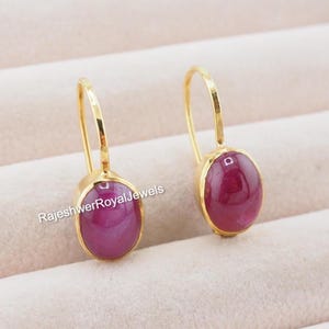 Puede incluir: Un par de pendientes dorados con piedras preciosas ovaladas de color rojo intenso. Las piedras preciosas están engastadas en un bisel dorado y los pendientes tienen un diseño de gancho simple. El texto "RajeshwerRoyalJewels" es visible.