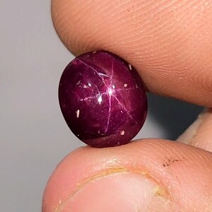 May include: A polished, oval ruby gemstone, held between fingers. The deep red gem has a star-like pattern across its surface, with small white inclusions. The gem is smooth and reflective, with a high lustre.